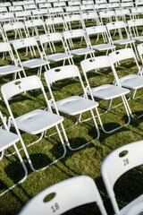 Empty folding chairs stand in rows on green grass, casting shadow on ground. Preparation for outdoor event, anticipation for guests to celebrate wedding, birthday banquet, solemn event party