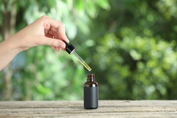 Wall Mural - Woman dripping tincture from pipette into bottle at wooden table against blurred green background, closeup