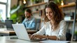© Yuliia - A smiling woman working on her laptop in a modern, collaborative office space, with a colleague in the background. Ideal for themes of teamwork, productivity, and a positive work environment.