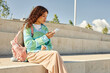 © Seventyfour - Side view of multiethnic curly girl with schoolbag dressed in pastel clothes using phone surfing net while sitting on concrete bench in park, copy space