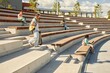 © Seventyfour - Wide angle shot of male student with skateboard and backpack walking down amphitheater stairs where teenagers sit and study after classes enjoying sunny day outdoors