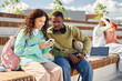 © Seventyfour - Smiling multiethnic schoolgirl engaged in watching funny videos on phone together with African American teenage boy while hanging out outdoors sitting on amphitheater bench
