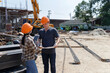 © skarie - Two construction workers discuss project details on-site, using a tablet and blueprints to plan the next steps amid the bustling construction environment.