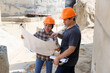 © skarie - Two construction workers discuss project details on-site, using a tablet and blueprints to plan the next steps amid the bustling construction environment.