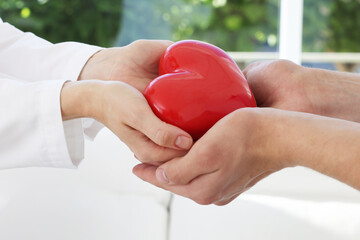 Wall Mural - Doctor giving red heart to patient at white table in clinic, closeup