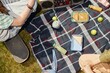 © Seventyfour - Top view of picnic cloth with snacks namely bottles of juice, sandwiches and apples on green lawn where unrecognizable teenagers sitting together and talking in park