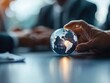 © praewpailyn - A close-up of a hand holding a glass globe, symbolizing global business and connection in a modern office environment.