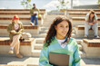 © Seventyfour - Portrait shot of multiethnic female student with curly hair posing in bright sunlight smiling at camera while standing on amphitheater steps outside, copy space