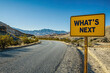 © Prime Lens - Under a clear sky, a yellow road sign asks 'WHAT'S NEXT?' as it stands before a winding desert road, symbolizing both uncertainty and future possibilities.