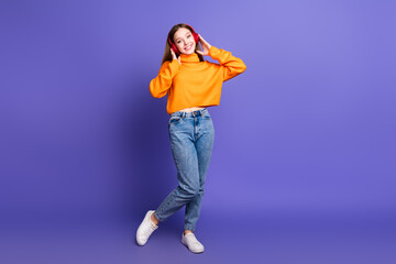 Wall Mural - Photo of blonde cheerful teenager girl in orange turtleneck and jeans hang out dancing listen headphones isolated on purple color background