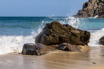 Waves crashing over a rock on the sandy beach at Portheras Cove, with a blue sky overhead