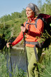 © pressmaster - Happy active woman with backpack taking walk with trekking sticks in natural environment while moving along riverside