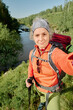 © pressmaster - Mature female backpacker in activewear looking at camera while taking selfie against riverside surrounded by green trees
