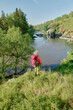 © pressmaster - Long shot of mature female backpacker in activewear standing on riverbank and looking at flow while admiring majestic landscape