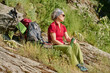 © pressmaster - Senior woman in activewear and sunglasses sitting on hill with green grass and having drink while admiring surrounding nature