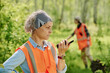 © pressmaster - Mature woman in headband and reflective vest reporting situation about accidents in the forest on walkie-talkie against another rescuer