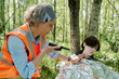 © pressmaster - Mature woman in safety vest checking pulse of young unconscious camper covered with foil while reporting about accident on radio set