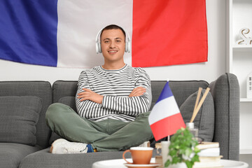 Canvas Print - Young man with headphones and flag of France sitting on sofa at home