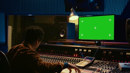 Wall Mural - Audio technician works on music post production with greenscreen in control room, pushing sliders and faders to adjust volume level. Producer mixing and mastering tracks with console in studio. Camera