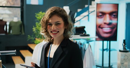 Canvas Print - Smiling retail clerk enjoys working in a fashion boutique, verifying stock on hangers and racks. Employee using tablet to check inventory items off of list, arranging clothes on discount. Camera B.