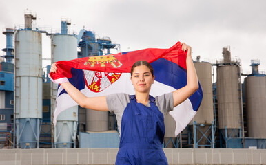 Smiling young woman in uniform posing with Serbia flag near factory