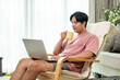© bongkarn - An Asian man is enjoying coffee while working on his laptop from a lounge chair in the living room.