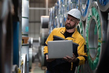 Metalwork manufacturing, warehouse for galvanized or metal sheet. Male engineer worker using laptop computer inspecting quality rolls of galvanized or metal sheet. Male worker working in warehouse