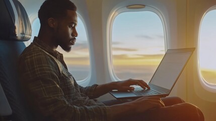 A man works on his laptop while sitting by the window of an airplane during a vibrant sunset