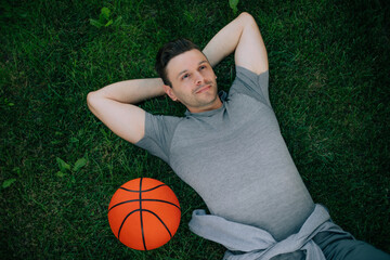 Wall Mural - A smiling man lies on a green lawn with his arms behind his head, next to a bright orange basketball. The relaxed pose and cheerful expression suggest a contented break after playing outdoors.