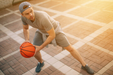 Wall Mural - A man in a backward cap and athletic wear is in a low, wide stance while holding a basketball, ready to make a move on the court. The golden sunlight casts a warm glow over the scene