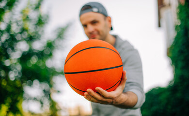 Wall Mural - A man in a gray hoodie holds a basketball close to the camera, with his face slightly blurred in the background. The outdoor setting features vibrant greenery, creating a fresh and energetic mood.