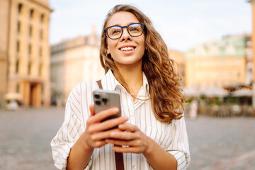 A young woman with curly hair and glasses thoughtfully using her smartphone in an urban plaza during the evening. Communication with friends. Technology, connection, blogging, modern lifestyle concept