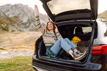 Wall Mural - A joyful young woman sitting by the open trunk of her car in a mountainous landscape during a sunny day, enjoying the great outdoors. Journey by car. 