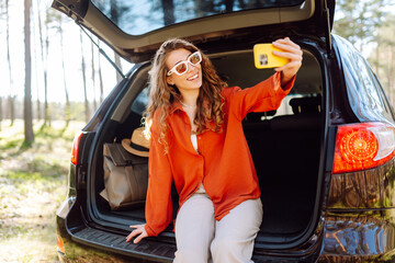 A cheerful woman sits on the trunk of her car in a vibrant forest and capturing a selfie with her smartphone, radiating happiness. Travel, weekend, nature, relax and lifestyle concept.