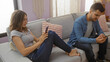 © Krakenimages.com - Couple sitting on couch in living room engaged on smartphones, man focused on phone in hand, woman leaning back on pillows, casual indoor setting