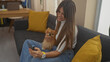 © Krakenimages.com - Smiling young woman holding a pomeranian dog in a cozy living room while using a smartphone.