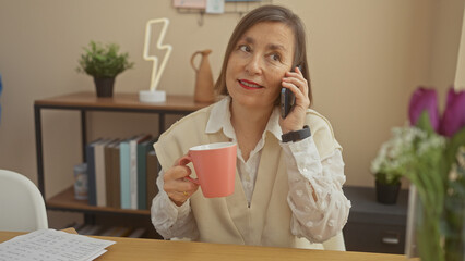 A mature woman enjoys a conversation on her phone while holding a pink mug in a cozy living room.