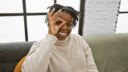 Poster - Smiling young black woman with dreadlocks making an okay sign indoors on a sofa, portraying a relaxed and playful mood.