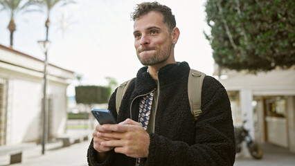 Sticker - Handsome young hispanic man with a beard using a smartphone on a sunny urban street, embodying a modern, connected lifestyle.