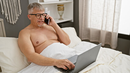 Poster - Grey-haired man using laptop and talking on phone in bedroom, showcasing remote work and modern lifestyle.