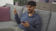 © Krakenimages.com - A smiling young man with a beard, using a smartphone, gestures while lounging on a sofa indoors.