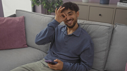 Wall Mural - A cheerful young man makes an ok gesture over his eye while holding a smartphone in a modern living room.