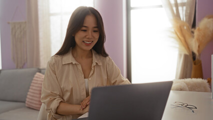 Canvas Print - Young chinese woman working on a laptop in a cozy indoor living room environment, showcasing a warm and inviting home setting.