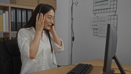 Asian woman wearing a headset and glasses, working on a computer in a modern office, showcasing a professional indoor workspace environment.