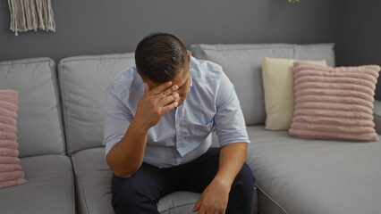 Canvas Print - Hispanic man feeling stressed sitting on a sofa in a living room with a gray interior and decorative pillows in an apartment.