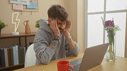 Wall Mural - A stressed hispanic man with a beard feels a headache at home while using a laptop near a vase with flowers.