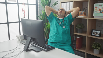 Hispanic male doctor in scrubs relaxing in hospital office with computer and stethoscope