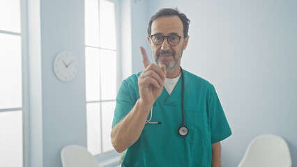 Poster - Hispanic male doctor in scrubs gesturing attention in a bright hospital room