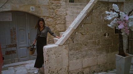 Wall Mural - A young beautiful hispanic woman walking in the charming streets of polignano a mare, puglia, italy, holding a basket under a stone staircase near a rustic building and flowery tree.