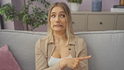 Wall Mural - Attractive blonde woman nervously pointing while sitting on a sofa in a cozy living room.